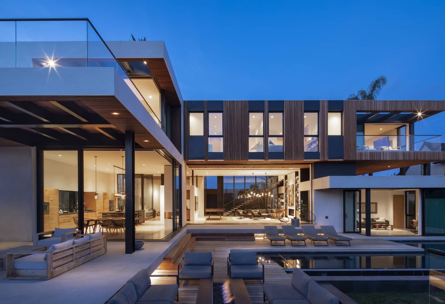 Modern residence at dusk: warm-lit interiors open to a pool deck, cantilevered wood-clad upper volume above